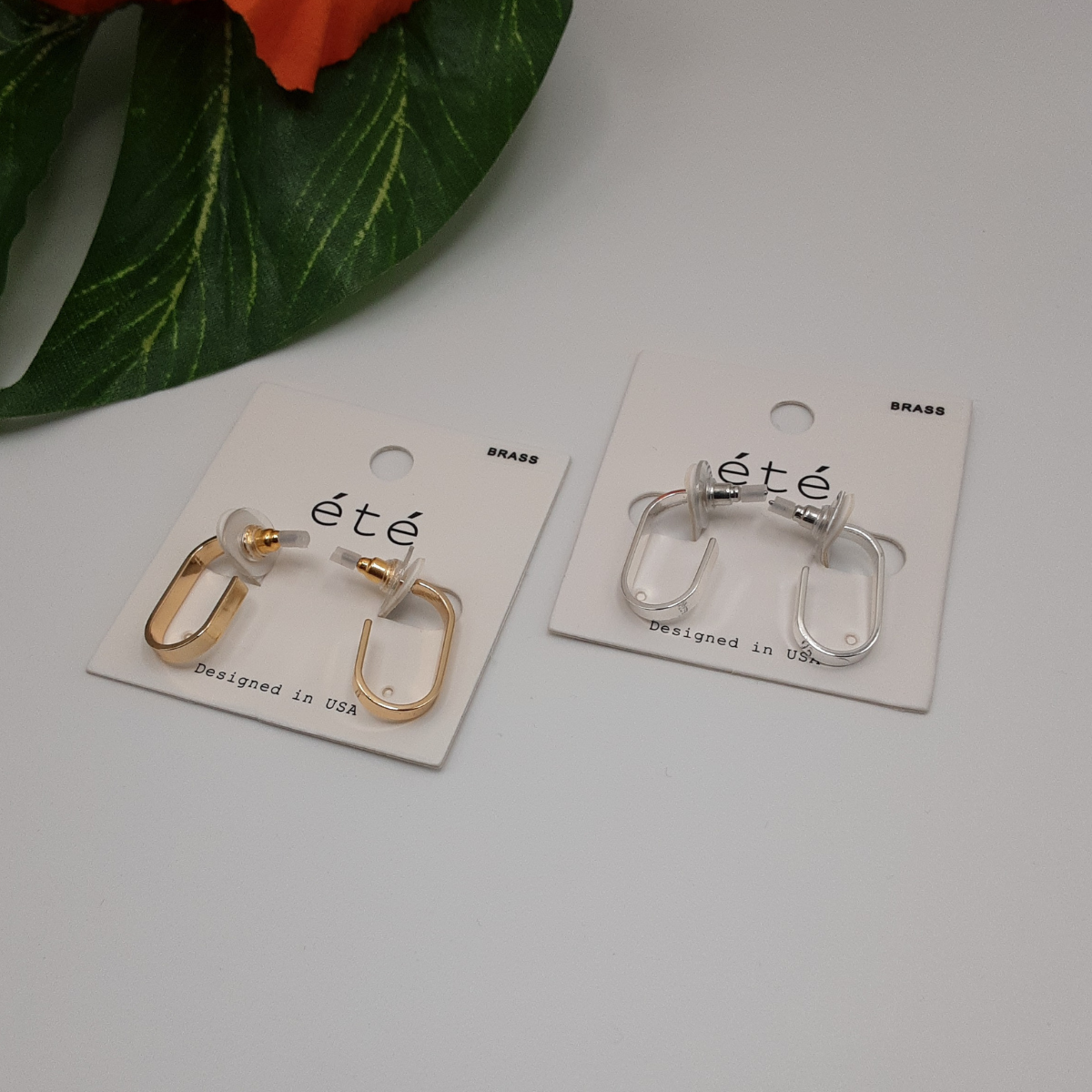 Two pairs of the J Hoop Earring from SPECIAL EFFECTS, one gold and one silver, are displayed on white cards labeled "été," set on a white surface with green leaves and orange flowers adorning the arrangement. Main image
