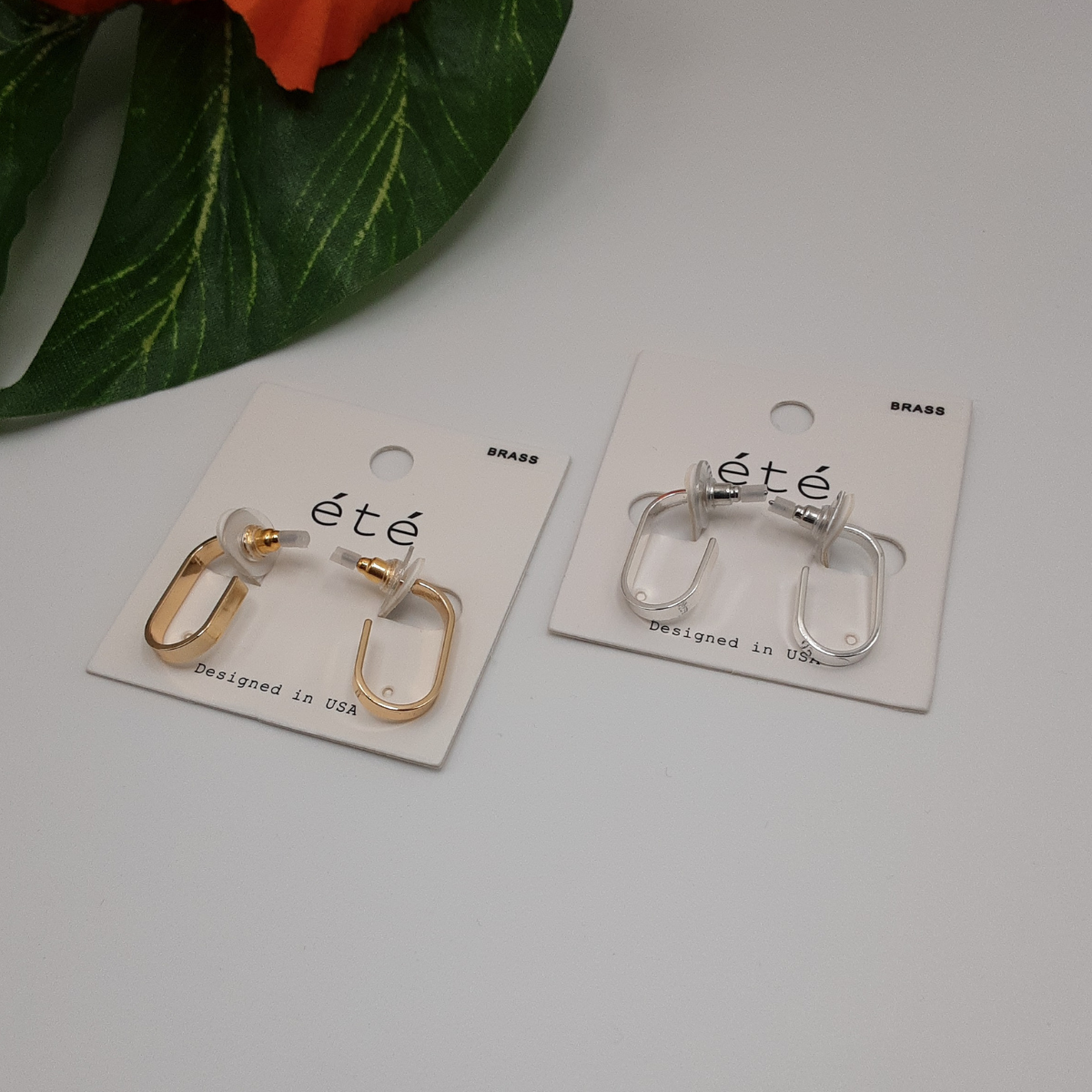 Two pairs of the J Hoop Earring from SPECIAL EFFECTS, one gold and one silver, are displayed on white cards labeled "été," set on a white surface with green leaves and orange flowers adorning the arrangement.