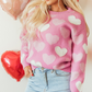 A person is wearing a Shewin 'Pearl Heart Print Valentines Crew Neck Sweater' paired with light blue jeans. The background features red and pink heart-shaped balloons, enhancing the charming scene.