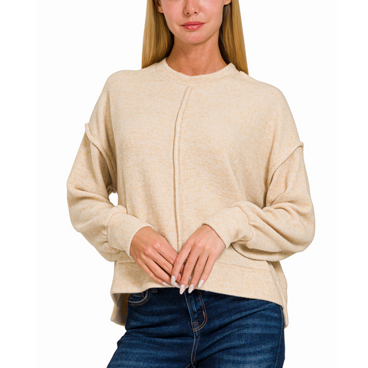 A person stands wearing the Brushed Hacci Hi Low Hem Sweater in Beige by Zenana and blue jeans against a white background.