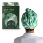 A person wearing the Plot Twist Turbo Towel by DM MERCHANDISING INC, featuring a green and white leaf pattern, enjoys hands-free drying with this microfiber hair towel. The matching towel packaging is displayed beside them.