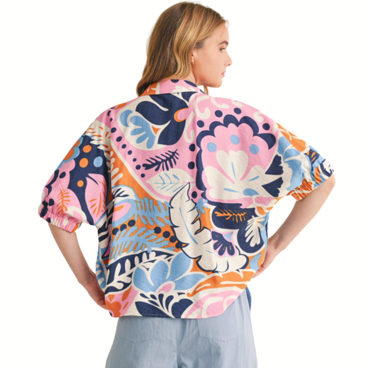 A blonde woman faces away, wearing the FASHION GO Multicolor Print 1/2 Sleeves Top with an open collared neck, loose fit, and an abstract floral pattern in pink, blue, orange, and white.