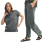 A woman models the Mono B Laguna Cowl Neck Wide Leg Pant Set—a matching dark gray lounge set with a cowl-neck short-sleeve top and wide leg drawstring pants, styled with black sandals against a white background.