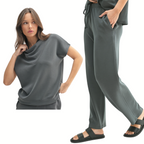 A woman models the Mono B Laguna Cowl Neck Wide Leg Pant Set—a matching dark gray lounge set with a cowl-neck short-sleeve top and wide leg drawstring pants, styled with black sandals against a white background.