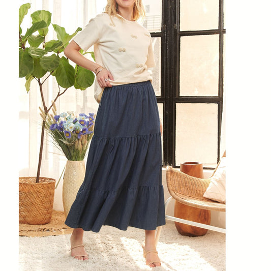 A woman stands indoors in a cream short-sleeve top and the FASHION GO Tiered Denim Maxi Skirt. Sunlight streams through the window, highlighting a large potted plant and a woven basket of flowers nearby.
