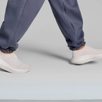 A person strolls in gray sweatpants and light gray slip-on shoes, exuding minimalist recovery. The OO My Zen in Chalk by OOFOS LLC features OOfoam™ technology, offering comfort and style against a plain backdrop.