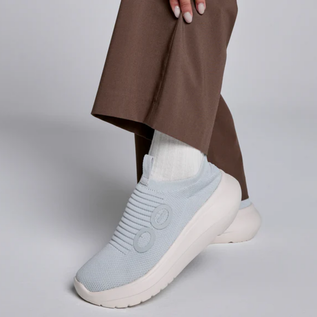 Wearing OOFOS LLC's OO My Zen in Sky, a light blue slip-on sneaker with a white sole and seamless circular knit design, someone pairs them with brown pants and white socks against a neutral background.