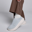 Wearing OOFOS LLC's OO My Zen in Sky, a light blue slip-on sneaker with a white sole and seamless circular knit design, someone pairs them with brown pants and white socks against a neutral background.