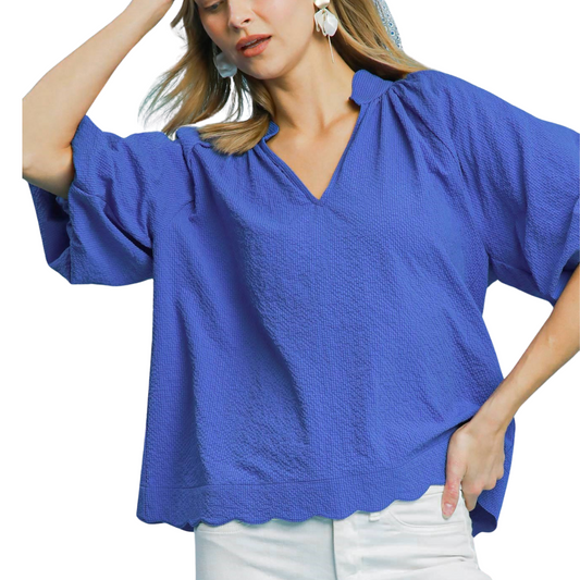 A woman wears the Umgee Bubbled Sleeved Seersucker Top in Ocean, featuring a loose fit, textured fabric, scalloped hemline, and elbow-length sleeves, paired with white pants.