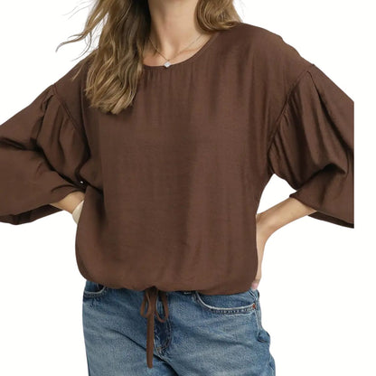 A woman wears the Umgee Round Neck Long Sleeve Drawstring Blouse, featuring a loose fit, adjustable drawstring hem, and wide sleeves, styled with blue jeans and her hands on her hips.