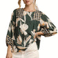 A woman wears the Umgee Mix Floral Print Blouse with Contrast Yoke, featuring green, white, and yellow patterns and long sleeves, paired with white pants for a relaxed look.