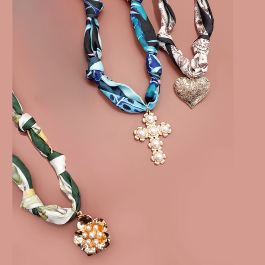 The FASHION GO Handmade Scarf Necklace comes in assorted colors/patterns, featuring unique charms like a pearl-studded flower, pearl cross, and ornate silver heart, creating a stylish accessory on a plain pink background.