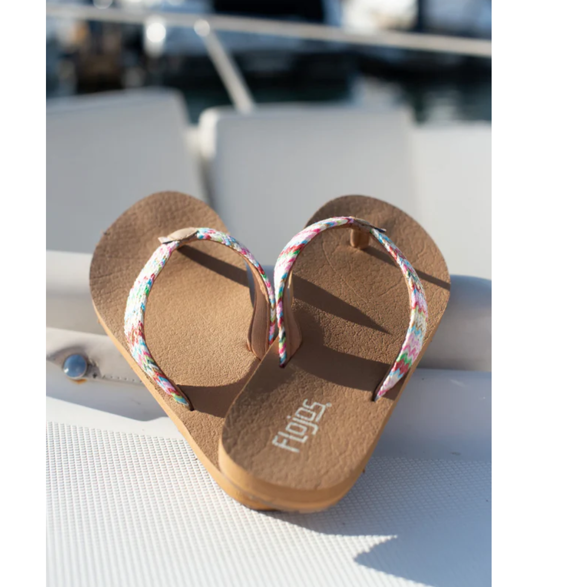 A pair of Juno Weave Flip Flops in Multi by Flojos sits on a light surface with water glistening behind. These FLOJOS sandals offer arch support and a yoga mat comfort footbed for delightful beach strolls.