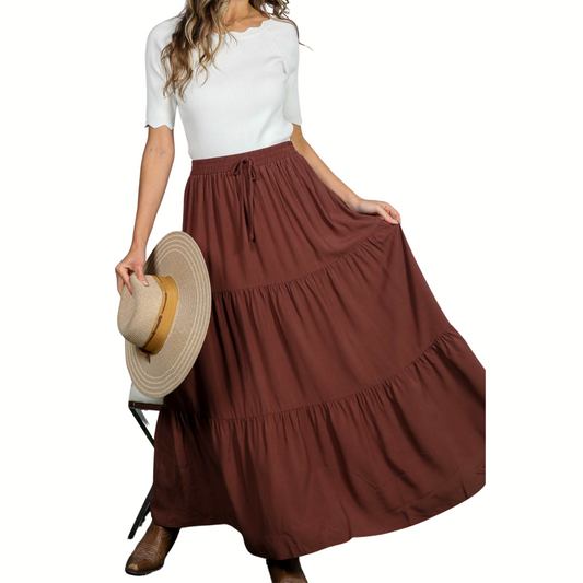 A person in a white short-sleeve top and Fashiongo Tiered Maxi Skirt stands wearing brown boots and holding a wide-brim hat.
