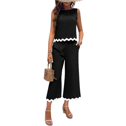 A woman in an elevated FASHION GO Two Piece Printed Sleeveless Pocket Set in Black, consisting of a black sleeveless top and matching black wide-leg pants with white wavy ribbon trim, is holding a wicker handbag. She is wearing sandals and has several bracelets on her wrist.