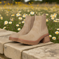 The Irie Block Heel Boot by MIA, crafted in tan suede with a contoured footbed, rests on a stone ledge with a field of daisies in the background.
