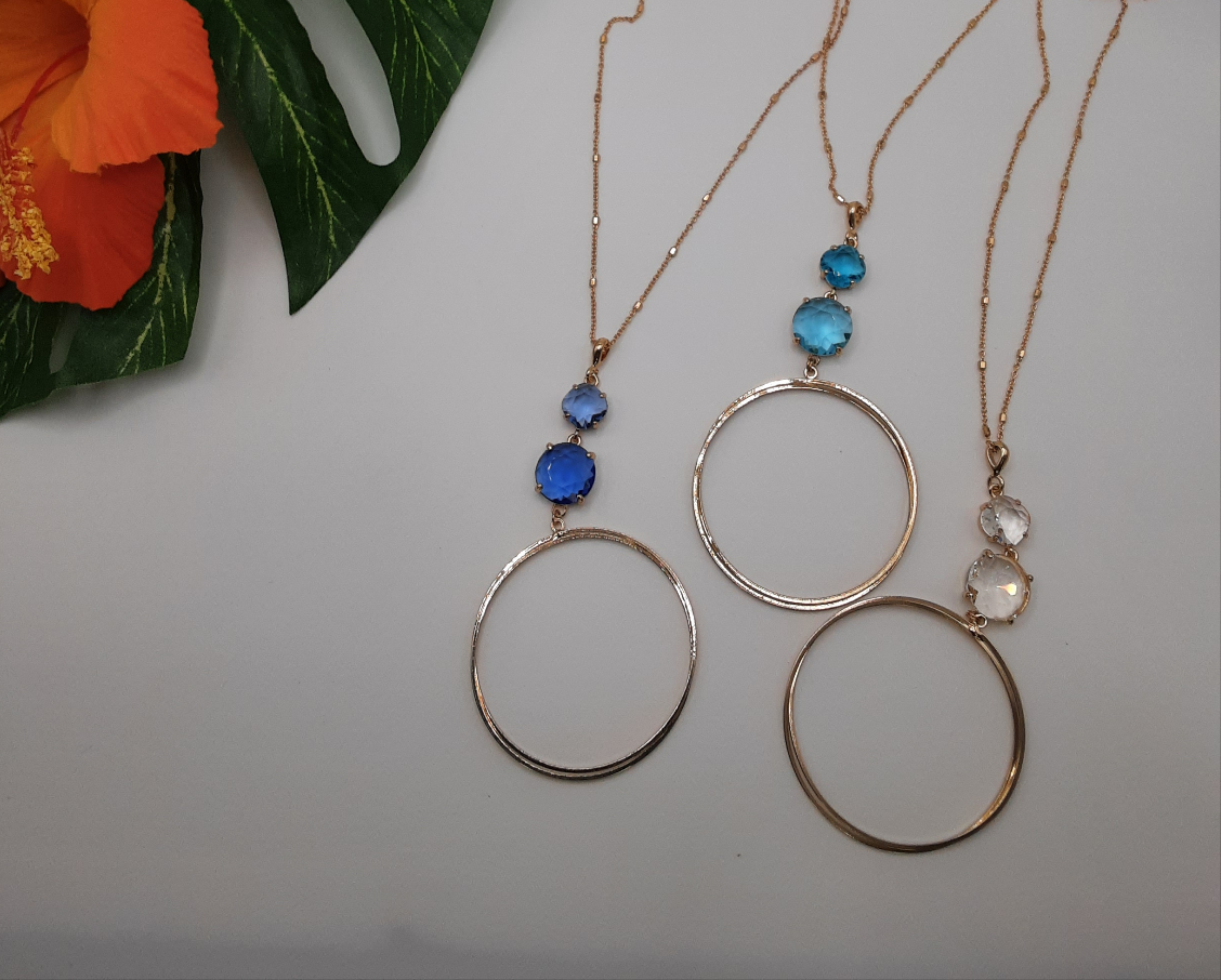 Three SPECIAL EFFECTS Crystal & Circle Long Pendants, gold necklaces each adorned with large circle crystal pendants featuring two gemstones in blue, teal, and white. On the left side of the image, a hibiscus flower and leaves enhance the scene with assorted colors. Main image