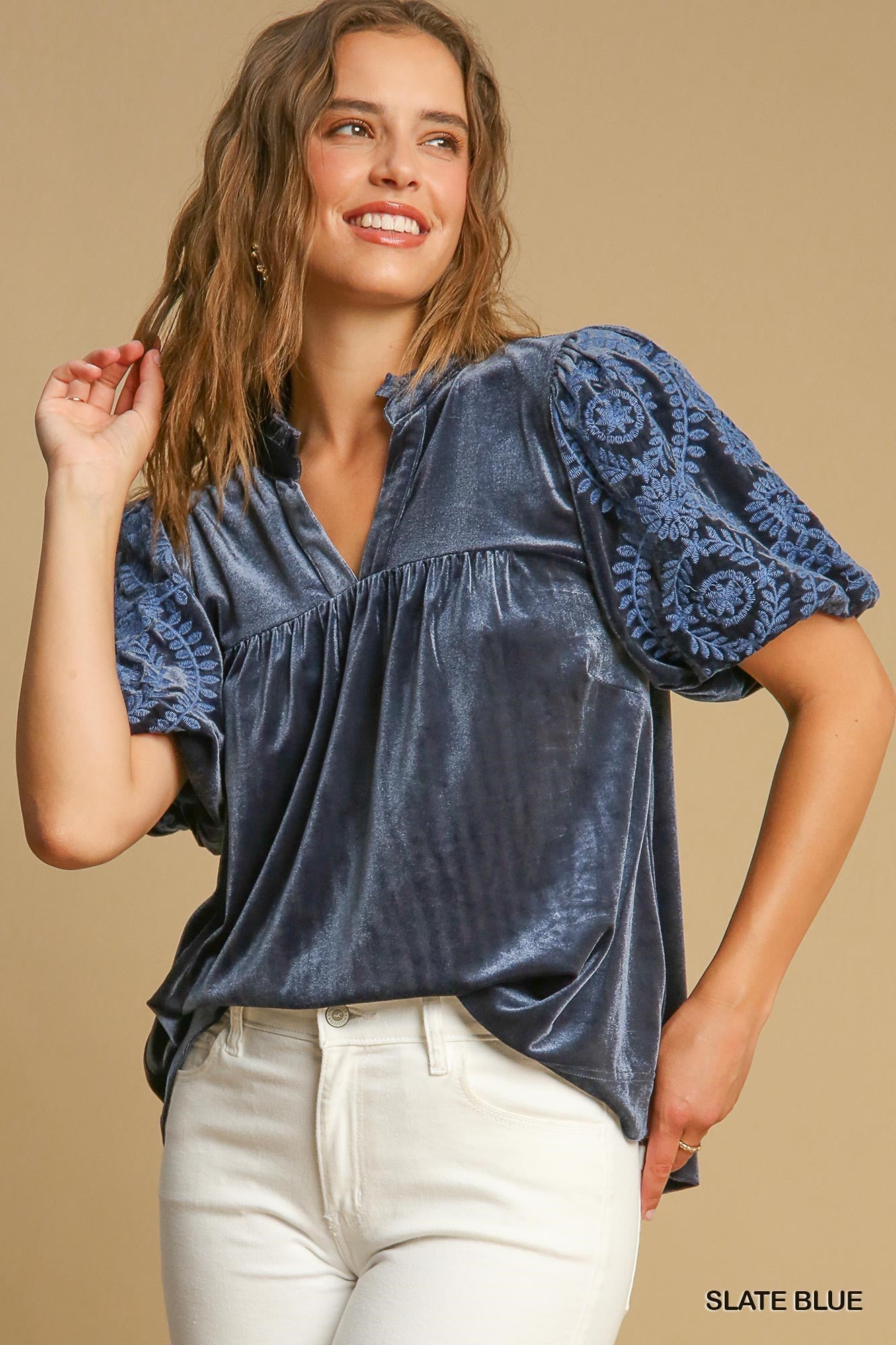 A woman wearing the Velvet Embroidered Sleeve Top in Slate Blue by Umgee, paired with white pants, smiles while posing against a beige background.