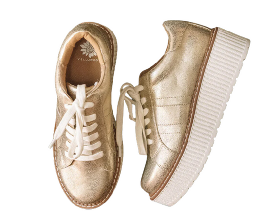 Marcell Casual Sneaker Tennis Shoe in Gold by Yellow Box features a pair of gold-colored sneakers with white laces and thick, white platform soles. The metallic uppers add a trendy touch, while the interior sole showcases a flower logo and the brand name "YELLOW BOX - CIT.