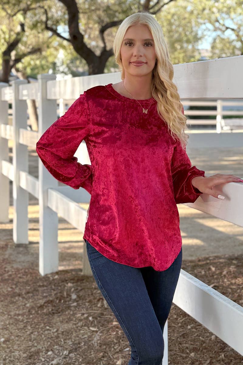 A woman in a 24-Hrs Fashion Crushed Velvet Top with flowy long sleeves and blue jeans stands by a white fence outdoors, trees behind her.