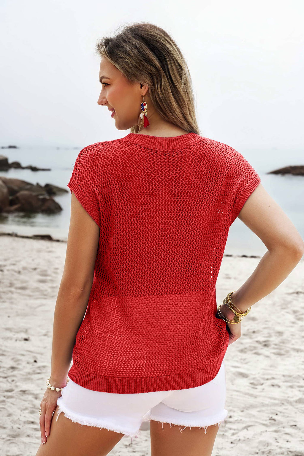 A woman stands on a sandy beach facing the ocean, wearing the 1 the Woman Flag Graphic Short Sleeve Sweater Top, paired with white shorts and jewelry.