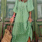 A woman in a Rosa Clothing Bohemian Floral Balloon Sleeves loose flare swing dress holds a woven straw bag, standing before rustic green wooden doors—an ideal look for a beach resort escape.