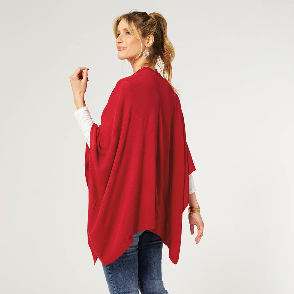 A woman stands in a three-quarter pose wearing the COCO + CARMEN Kiara Lightweight Wrap in red over a white long-sleeve top and blue jeans, set against a plain light background.