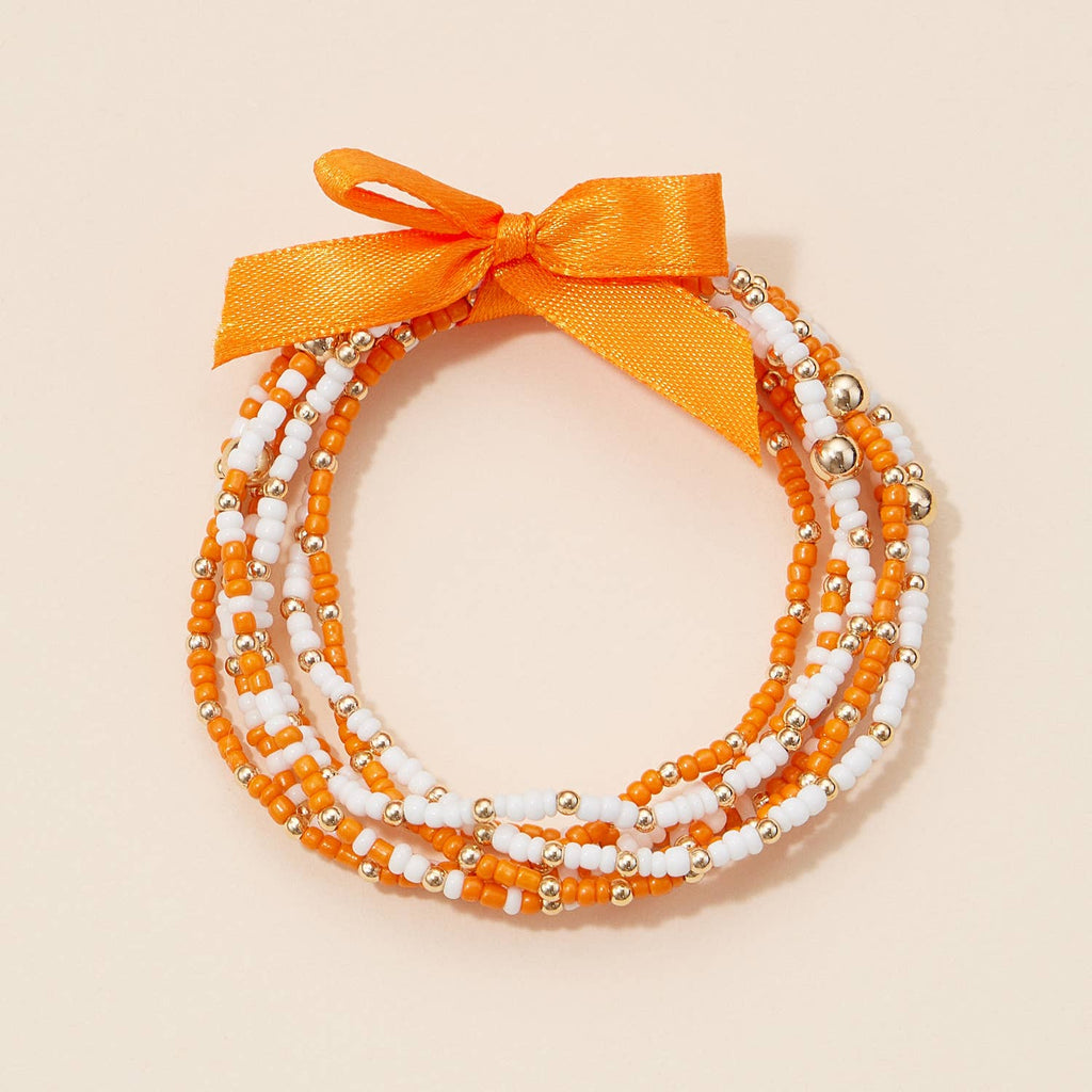 A Game Day Seed Beaded Bracelet Set consisting of four colorful seed beaded bracelets in orange, white, and metallic gold, tied together with an orange ribbon bow.