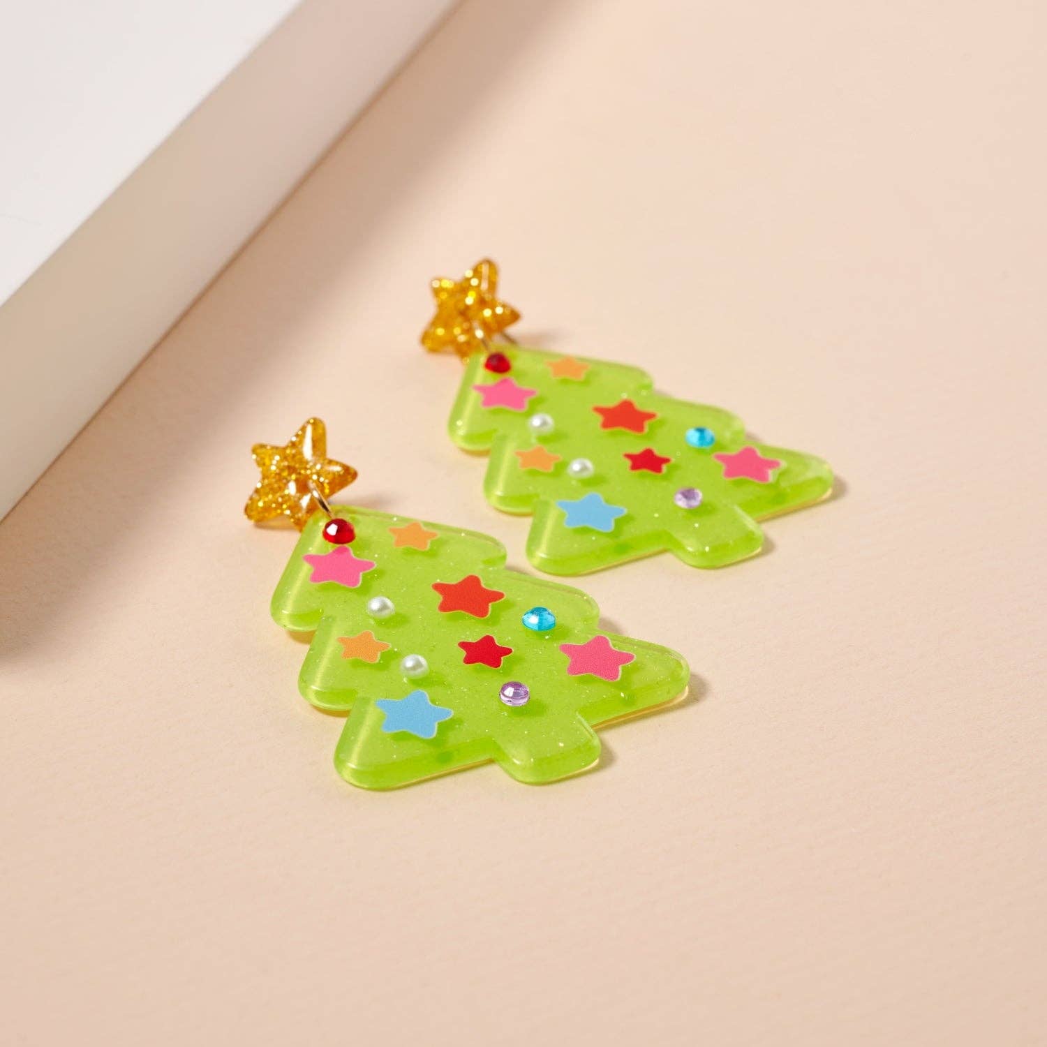 Show off your holiday spirit with the Glittery Christmas Tree Drop Earrings from Lilla Haven—these festive acrylic earrings feature vibrant green trees adorned with colorful stars and dots, topped with sparkling gold stars for a whimsical touch. Perfect statement jewelry for the season, they’re stylishly displayed on a light backdrop.
