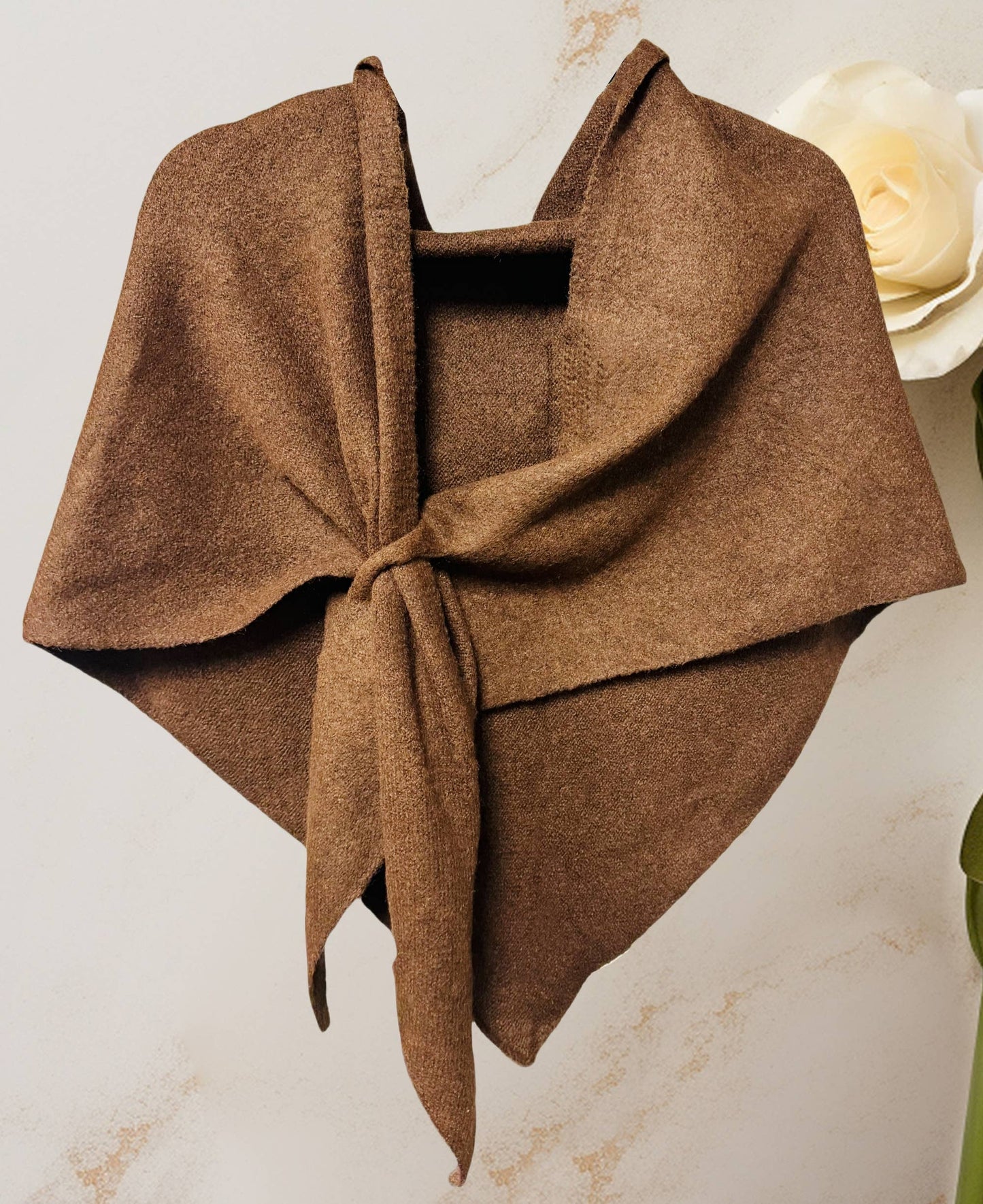 The JC Sunny Fashion Cashmere-Blend Knit Triangle Shawl with Cross-Front Design, in brown, is tied loosely and displayed against a light background with a white artificial rose partially visible on the right.