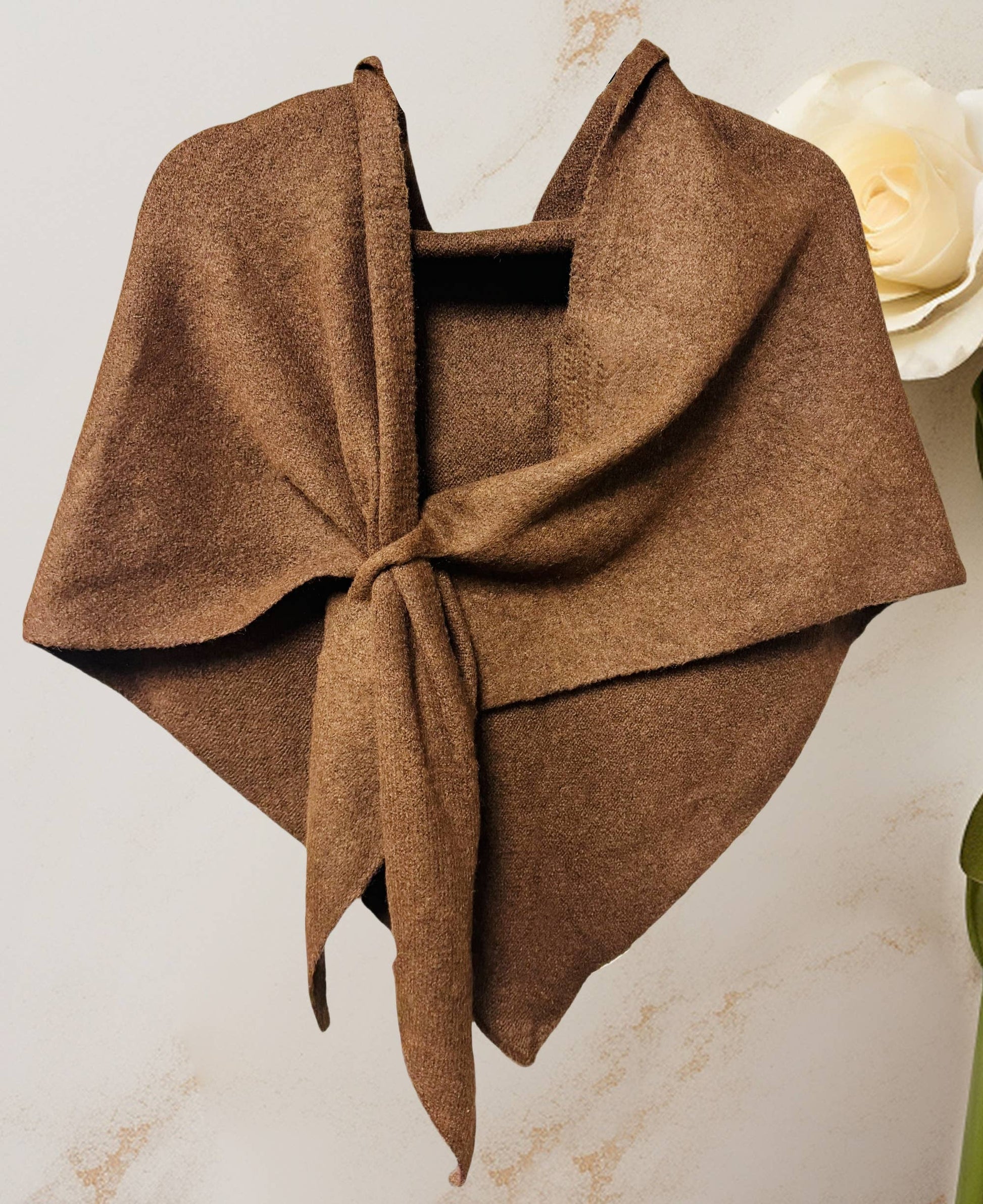 The JC Sunny Fashion Cashmere-Blend Knit Triangle Shawl with Cross-Front Design, in brown, is tied loosely and displayed against a light background with a white artificial rose partially visible on the right.