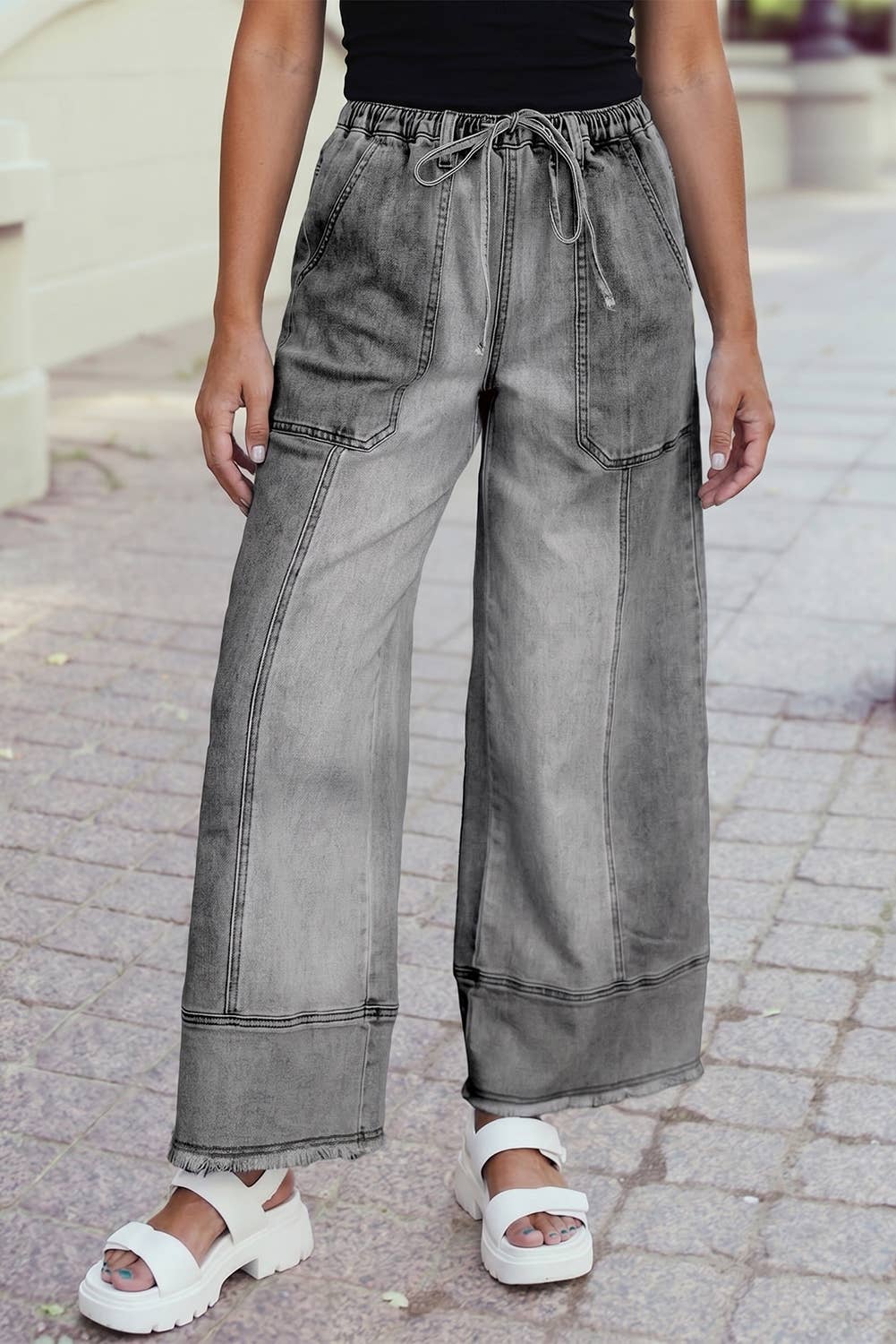 A person in a black top, 1 the Woman Drawstring Seam Detail Raw Hem Wide Leg Denim Pants, and white platform sandals stands on a paved sidewalk.