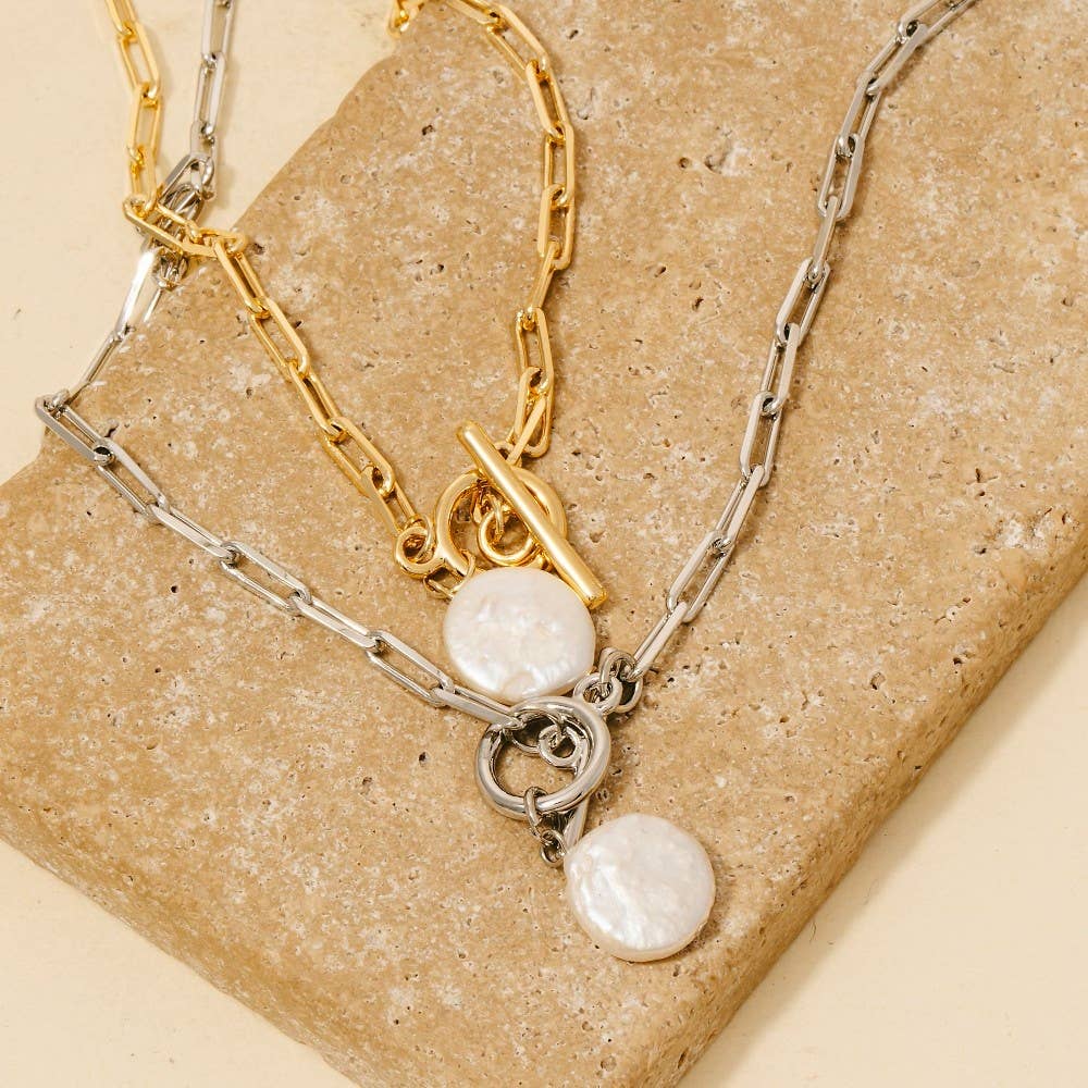 Two Anarchy Street Toggle Chain Link Disc Pearl Charm Necklaces, one gold and one silver, each with a round white disc pendant, are displayed on a rectangular beige stone surface.