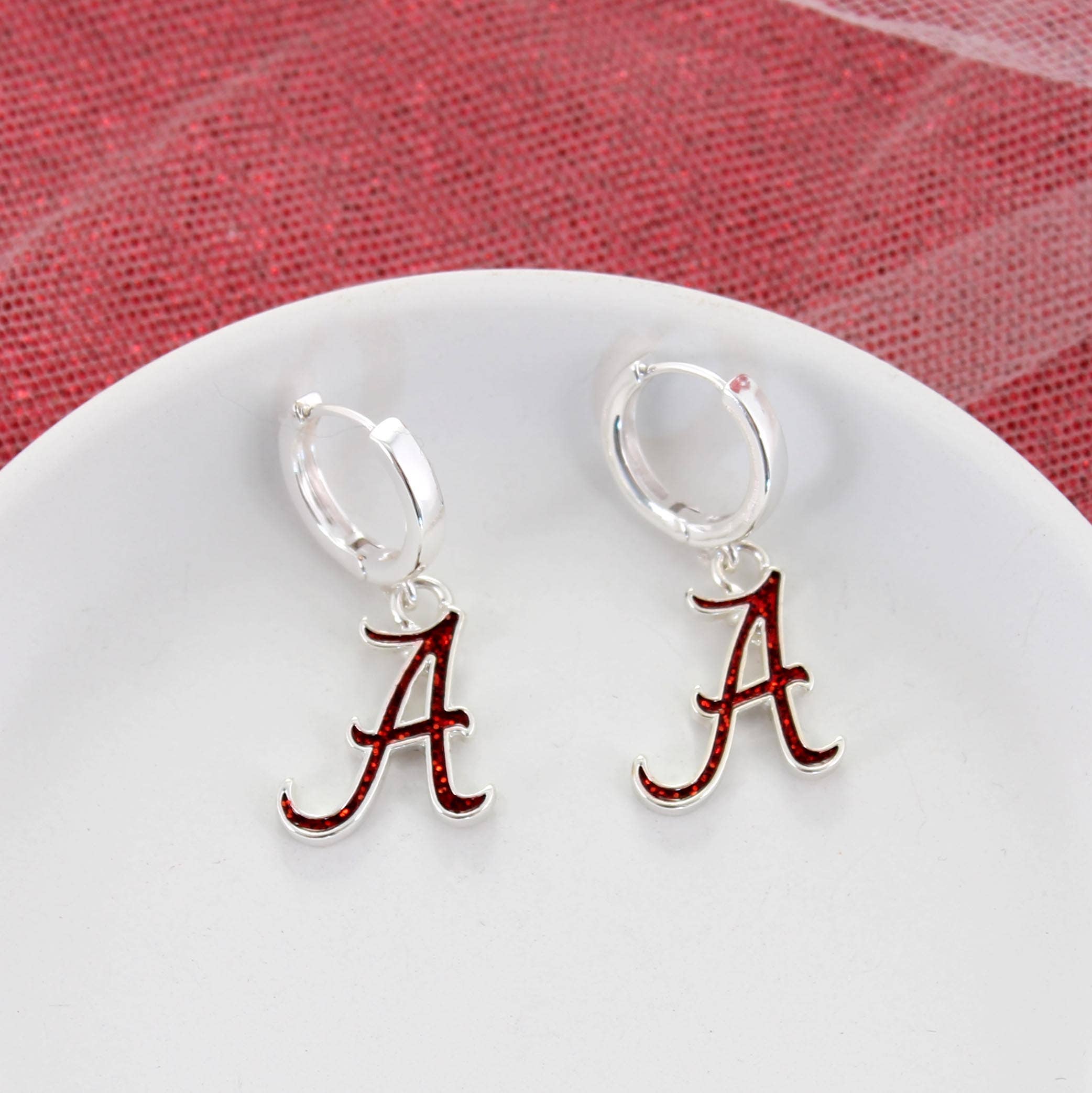Seasons Jewelry’s Alabama Glitter Enamel Logo Huggie Hoop Earrings—silver hoops adorned with red and silver letter "A" charms and sparkling crimson glitter enamel—are showcased on a white plate against a textured red and white backdrop. Main image