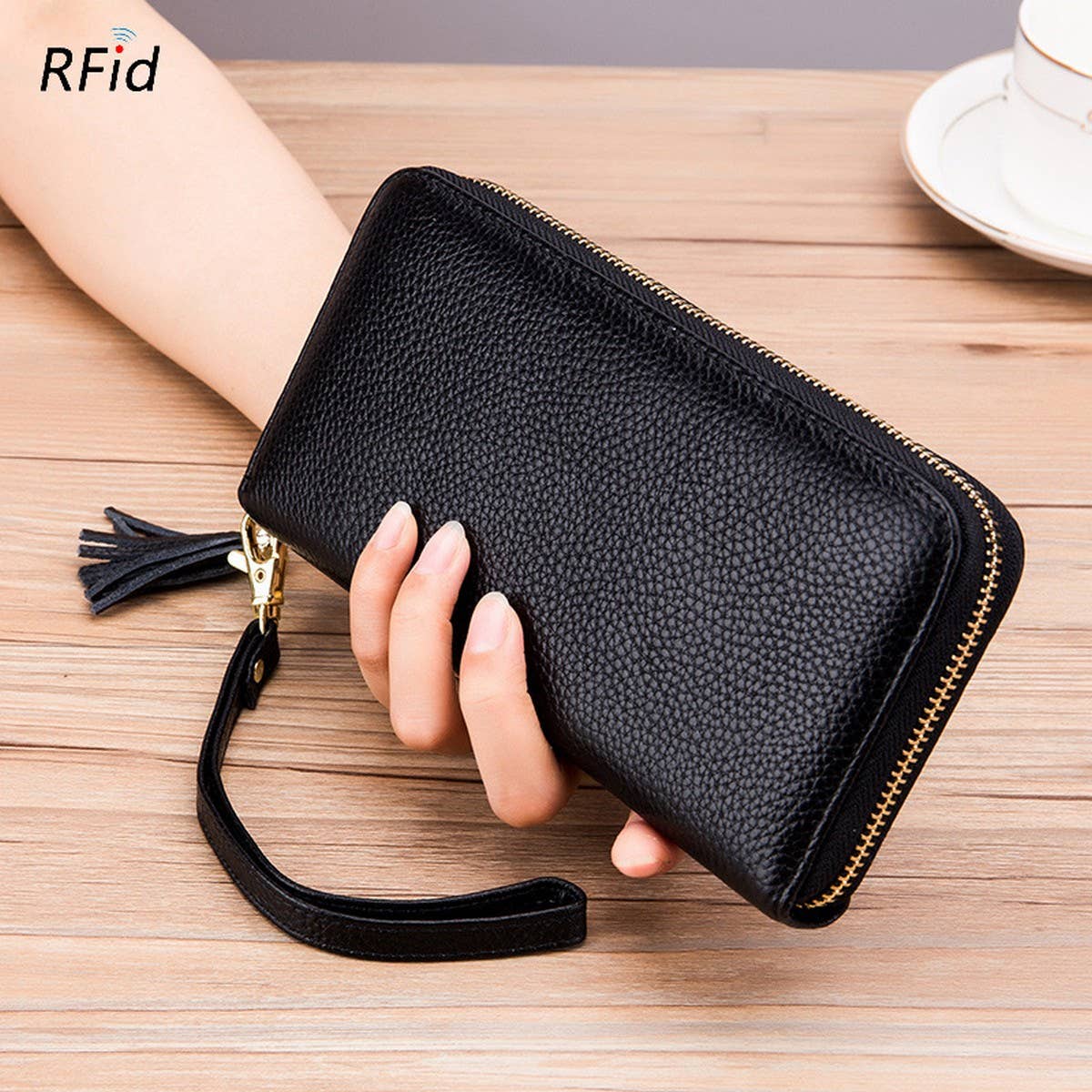 A hand holds the ACCITY ZIPPER TASSEL ANTI-THEFT BRUSH LEATHER WALLET, featuring black textured leather, a zipper tassel, and wrist strap, placed on a wooden table beside a cup and saucer.