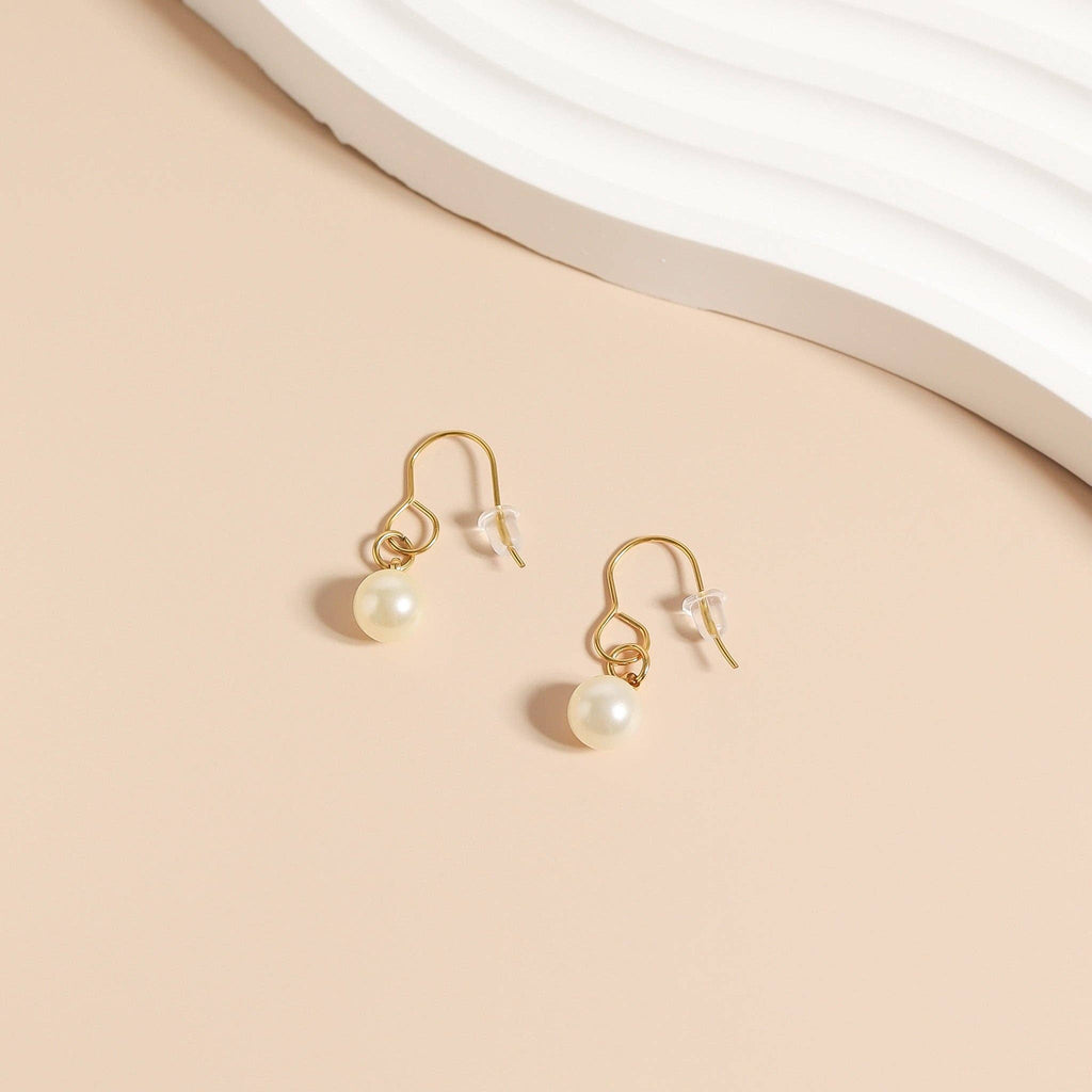 A pair of WJW Silicone Earring Backs (1 PAIR) is shown with gold hook earrings featuring white pearl drops, displayed on a light beige surface with a wavy white accent.