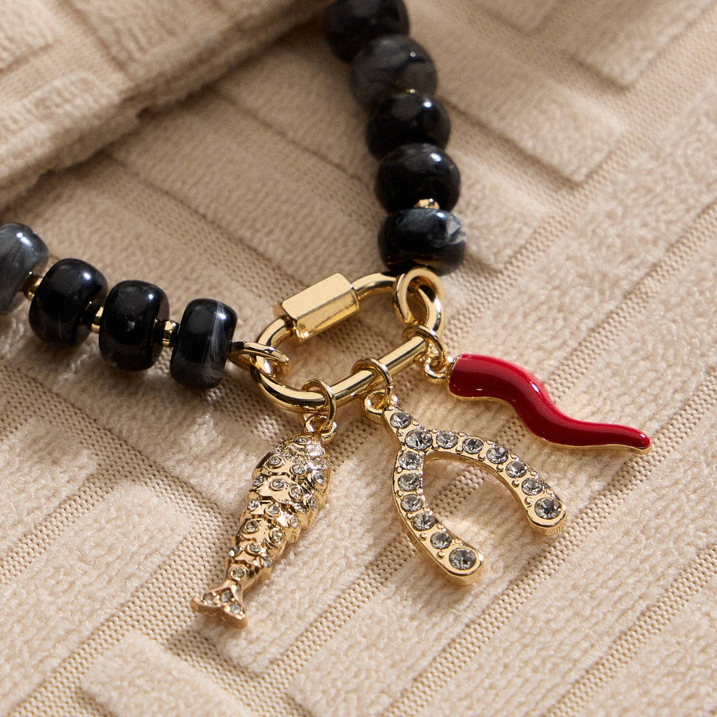 A close-up of the Lilla Haven Textured Glass Bead Necklace with Charm Trio showcases its black textured glass beads, gold-toned spacers, and three gold charms—a fish, a rhinestone-studded wishbone, and a red chili pepper—resting on beige textured fabric.
