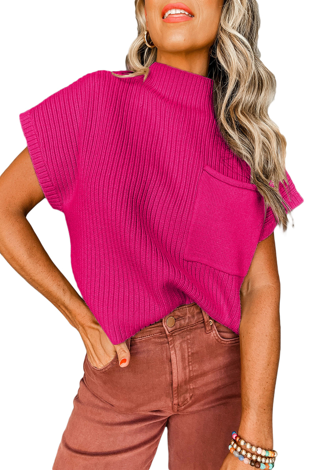 A person wears the Patch Pocket Ribbed Knit Short Sleeve Sweater by Shewin in vibrant pink, paired with brown pants. Their wavy hair and colorful bracelets make it ideal for transitional weather.