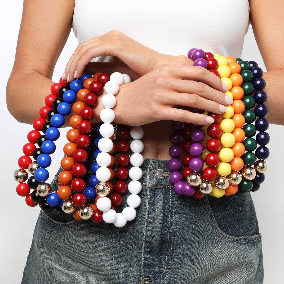 A person in a white top and denim skirt holds multiple colorful beaded necklaces, including the Marjorie Game Day Beaded Necklace by Canvas Style, featuring chunky oversized beads in red, blue, orange, white, purple, yellow, and green strands accented with metallic beads.