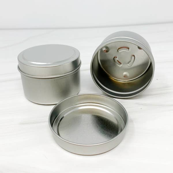 Three 4oz Mahogany Teakwood candles in round metal tins from Stephanie Henry Candle Company. One tin is upright and open, showcasing an interior with a hole pattern for soy wax candles, while the others are closed on a white surface.