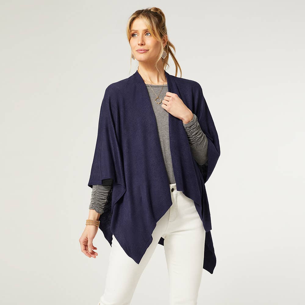 A woman faces slightly left against a light background, wearing the COCO + CARMEN Kiara Lightweight Wrap in navy blue over a gray top and white pants.