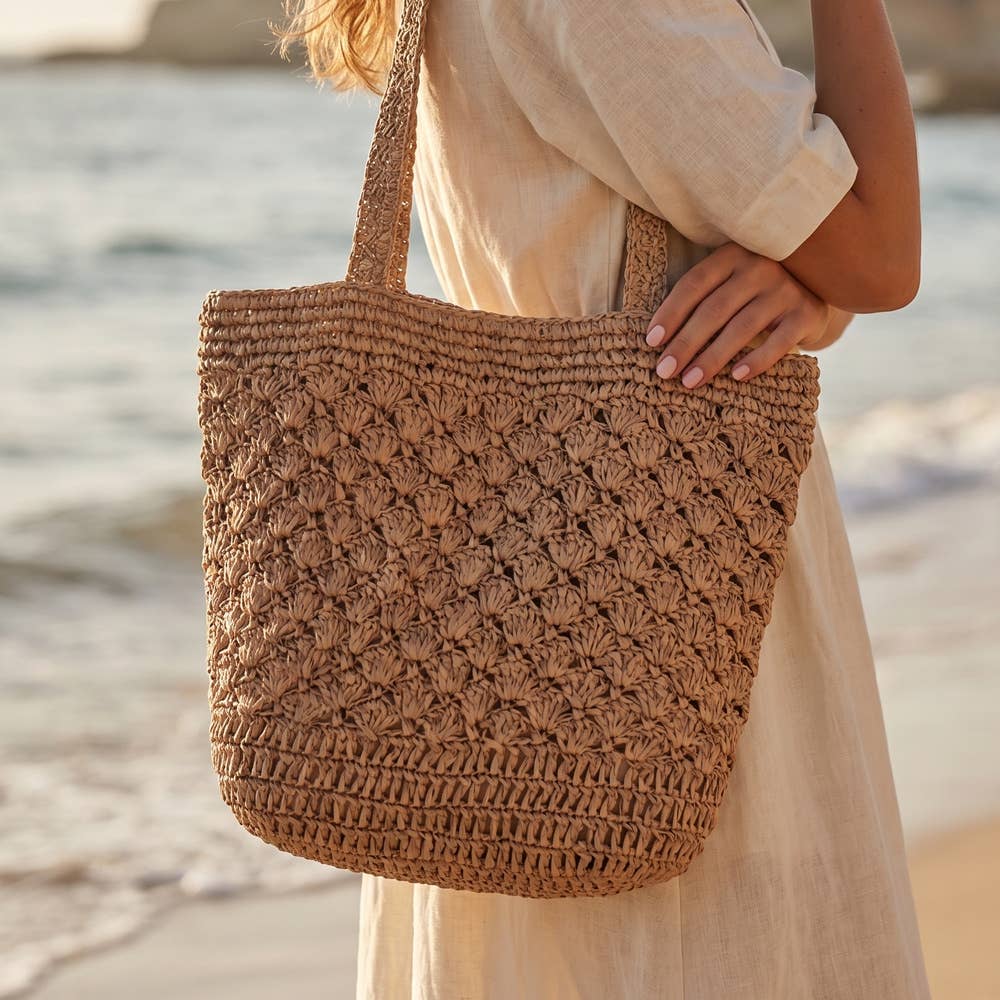 A person in a light-colored dress stands on the beach, holding the Anarchy Street Large Woven Straw Fan Stitch Shoulder Tote Bag.