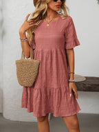 A woman stands indoors against a white wall, wearing the Suhkasana Loose Casual Short-Sleeve Dress in mauve, styled with sunglasses, bracelets, a necklace, and a straw handbag.