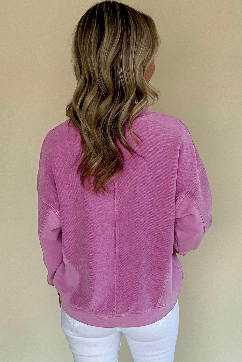 A person with long, wavy hair is turned away, wearing a Shewin Notched Neck Exposed Seam Drop Shoulder pink sweatshirt and white pants against a light-colored background.