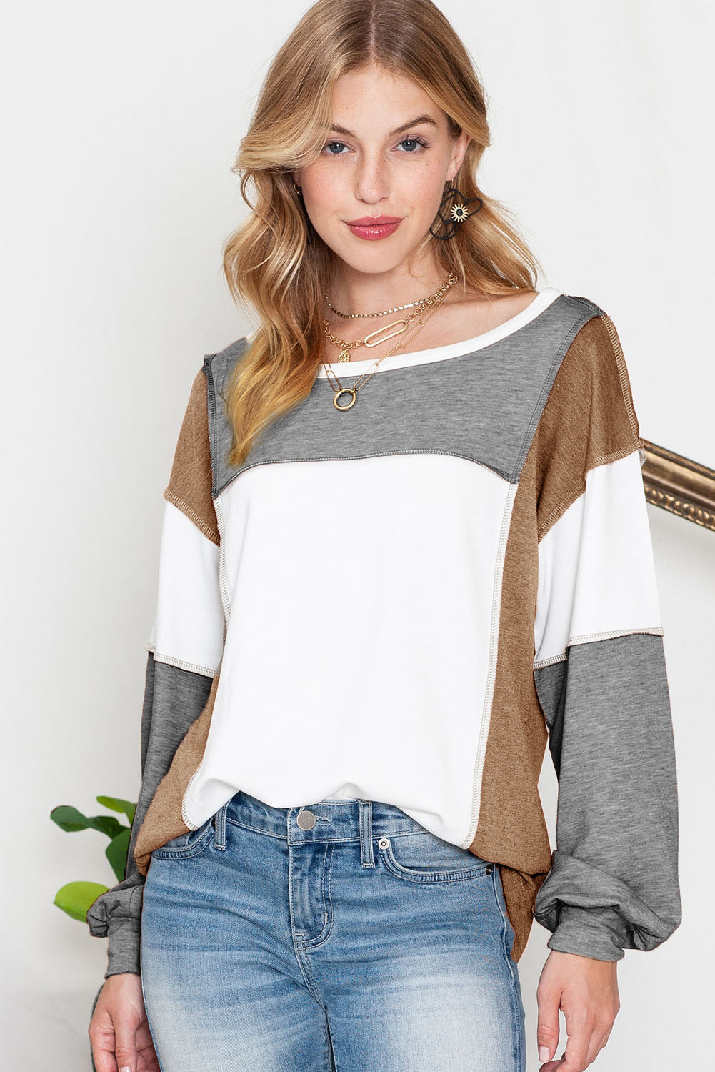 A woman with blonde hair is wearing the Shewin Khaki Color Block Exposed Seam Long Sleeve Top, featuring sections of gray, white, and versatile khaki. She has paired it with light blue jeans and is standing indoors against a light background.