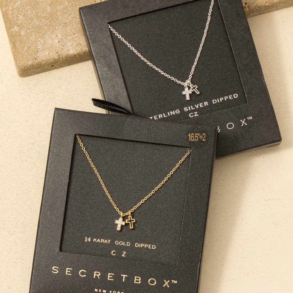 The Anarchy Street Secret Box Gold Dipped Cz Cross Pendant Necklace features two small cross pendants and is shown in both silver and gold finishes, displayed on a beige surface.
