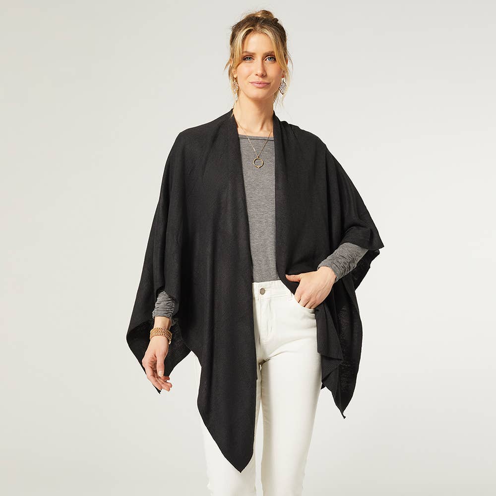 A person wears a black COCO + CARMEN Kiara Lightweight Wrap over a grey top and white pants against a plain light background. This one-size versatile piece brings effortless style and comfort to any outfit.