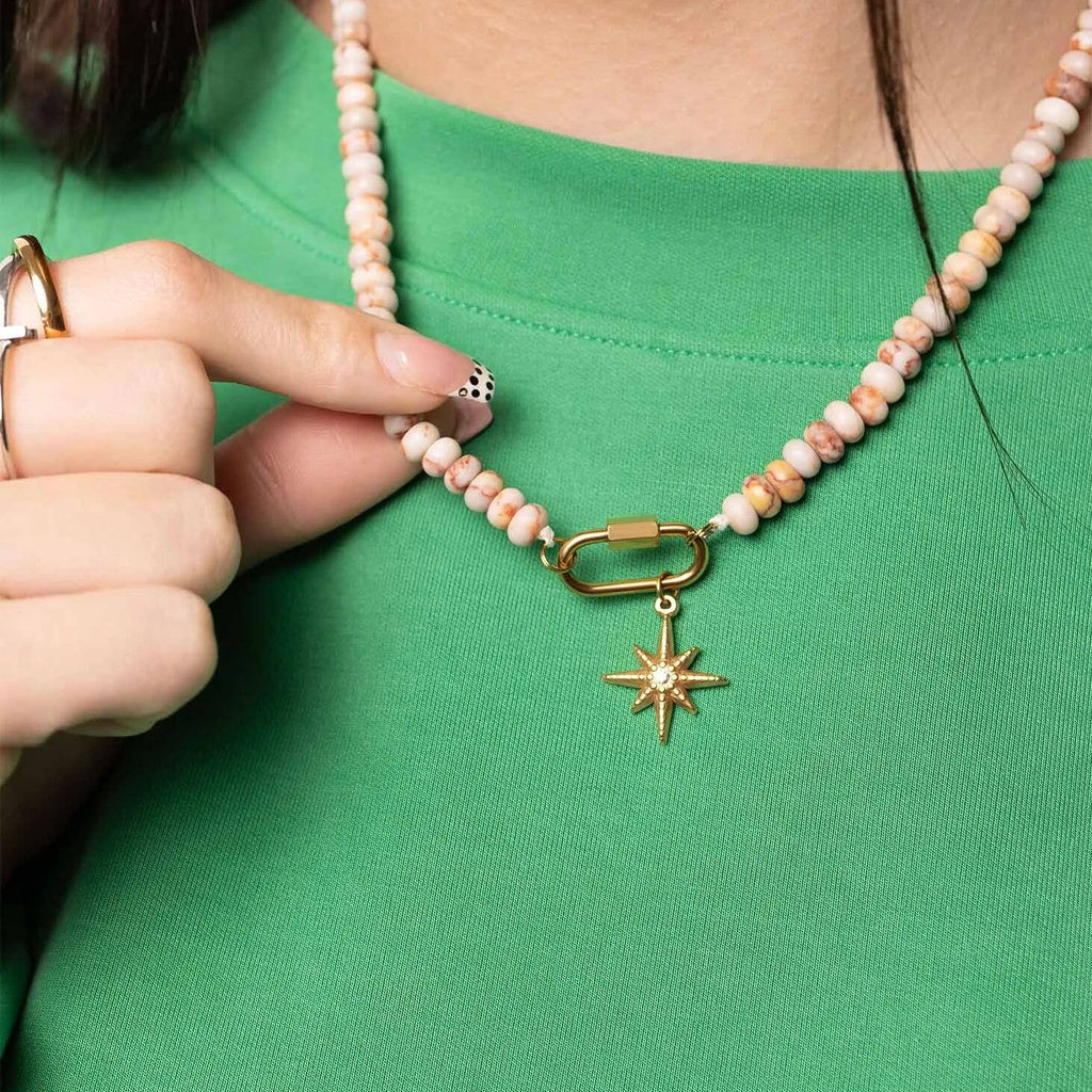 A person wearing a green shirt holds the WJW Stainless Steel Chunky Glass Bead Carabiner Necklace, which is adorned with a gold star pendant. Their fingernails display a black and white dotted design.