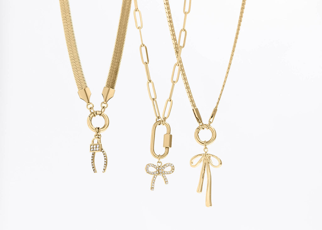 Three gold necklaces by WJW displayed on a white background: one featuring a clasp with crystal detailing, one adorned with a crystal bow charm, and the 18K Gold PVD Paperclip Carabiner Necklace showcasing a sleek gold bow charm.