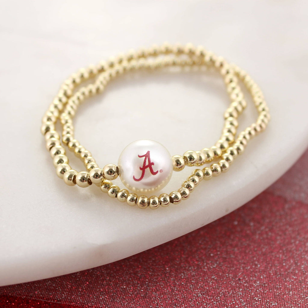 Seasons Jewelry's Alabama Gold & Pearl Logo Stretch Bracelet Set features gold beading and a white pearl with a red "A" Bama logo, elegantly displayed on a white marble surface—a stylish University of Alabama accessory perfect for any fan.